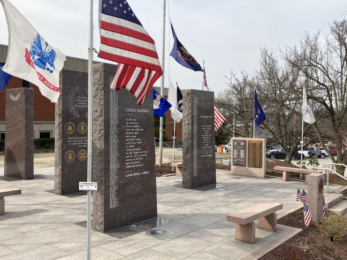 Memorial plaza with engraved stone pillars, U.S. and blue flags, and a bench under bare trees.
