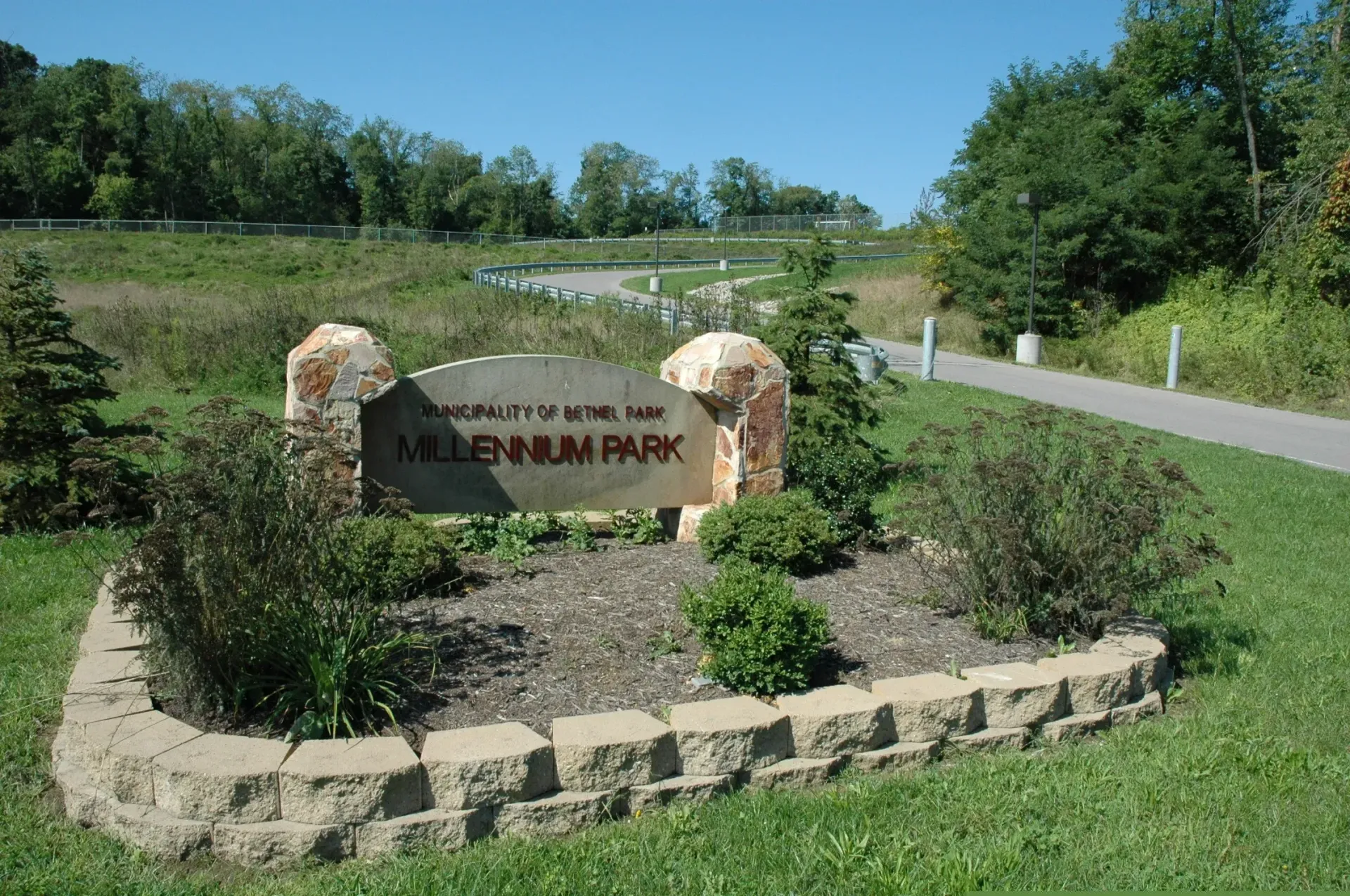 Stone entrance sign in a grassy park beside a paved path, with flowers and trees under a blue sky.