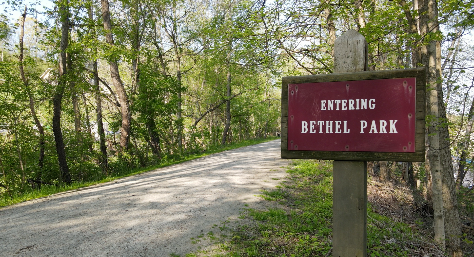 Dirt road entrance to Bethel Park with a red sign beside trees and greenery