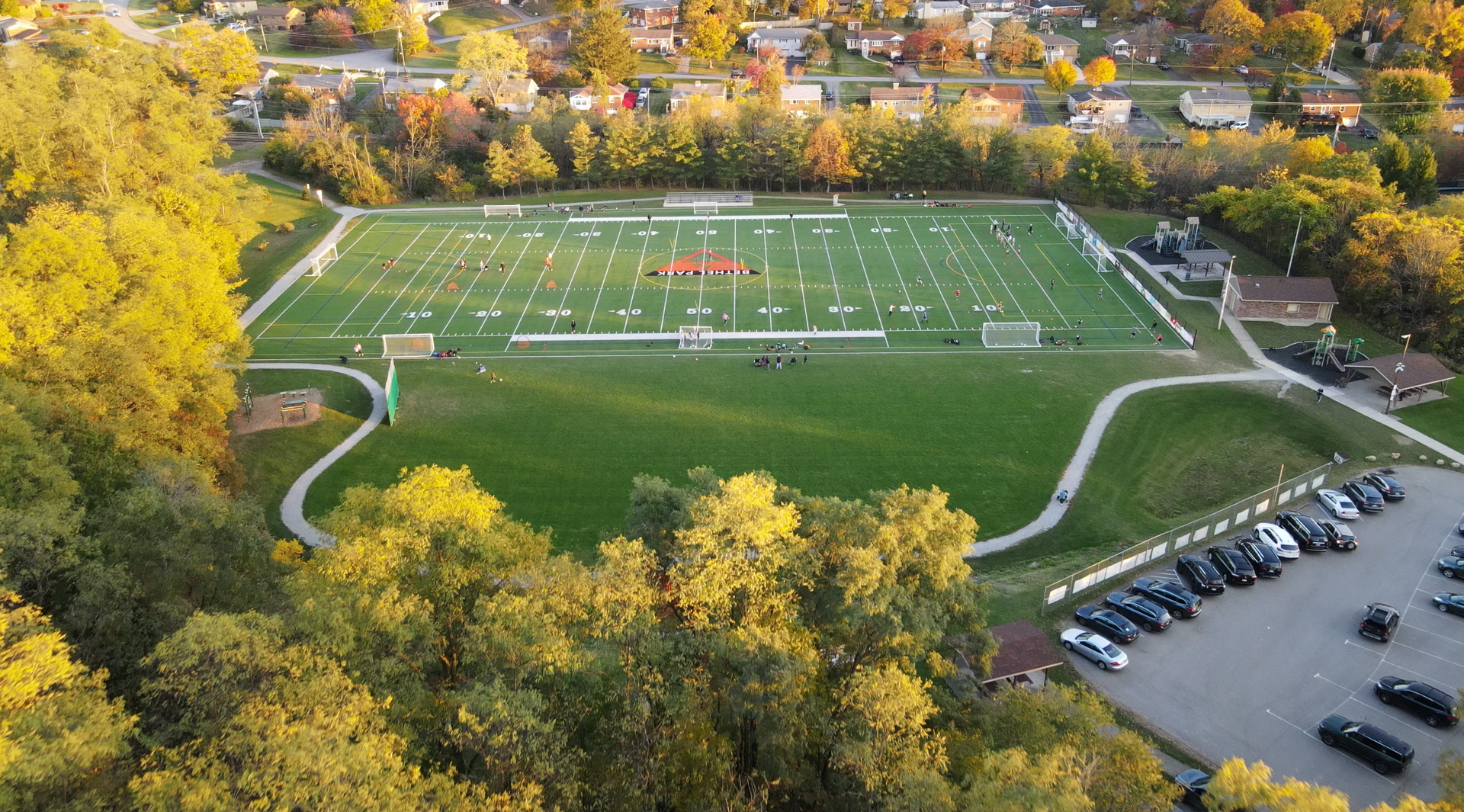 Aerial view of a lit outdoor football field surrounded by trees and a parking lot in autumn.
