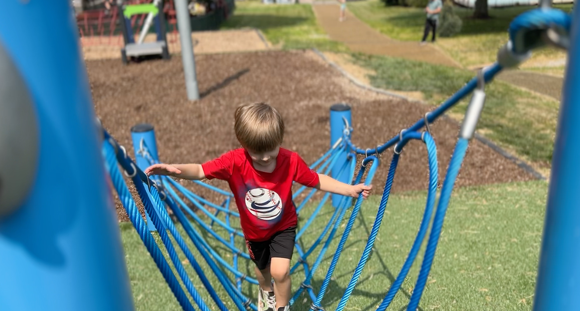 Child crossing a blue rope bridge at a playground, wearing a red shirt and black shorts