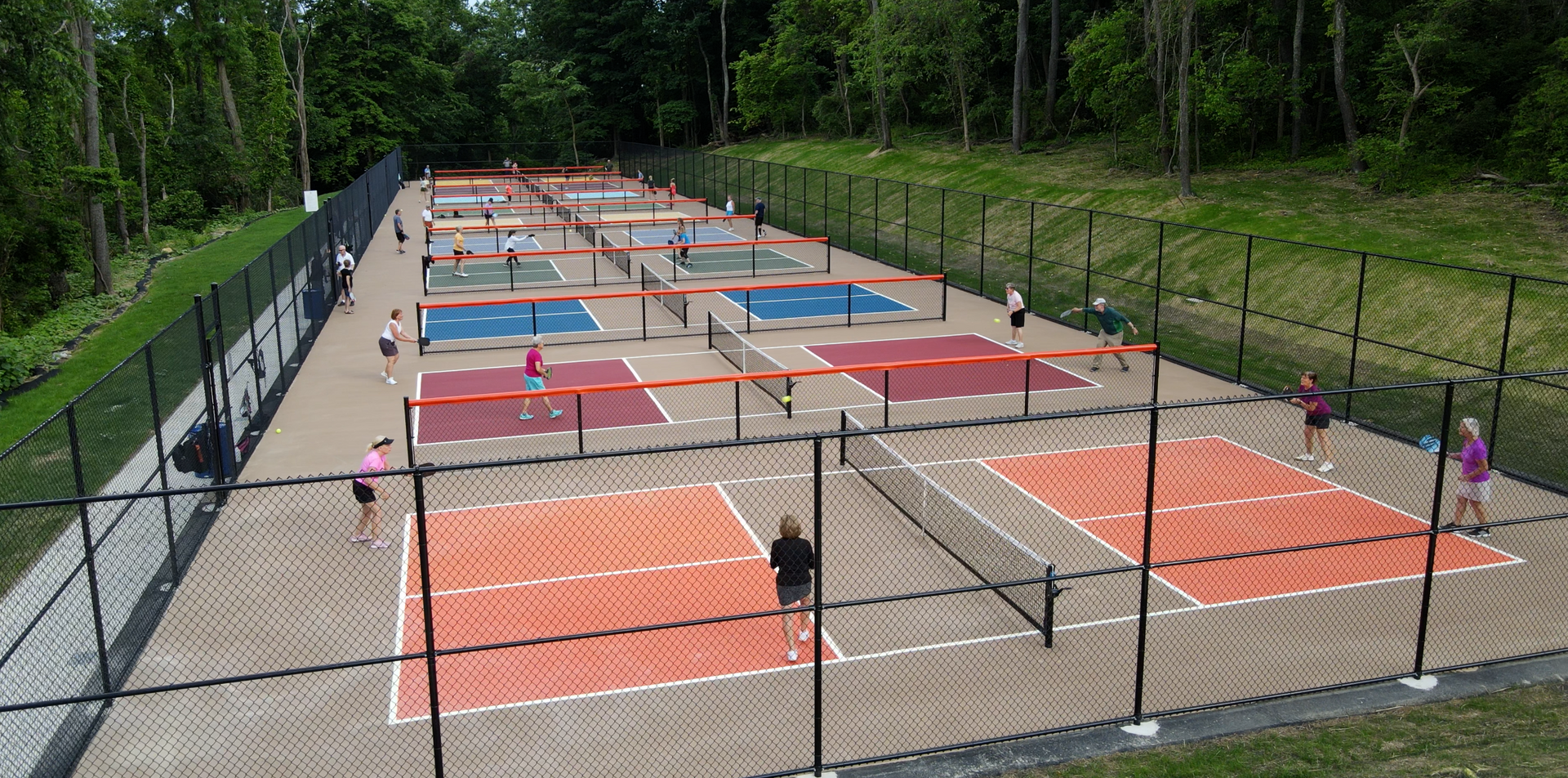 Outdoor tennis courts with several people playing on red and blue surfaces, surrounded by trees and fencing
