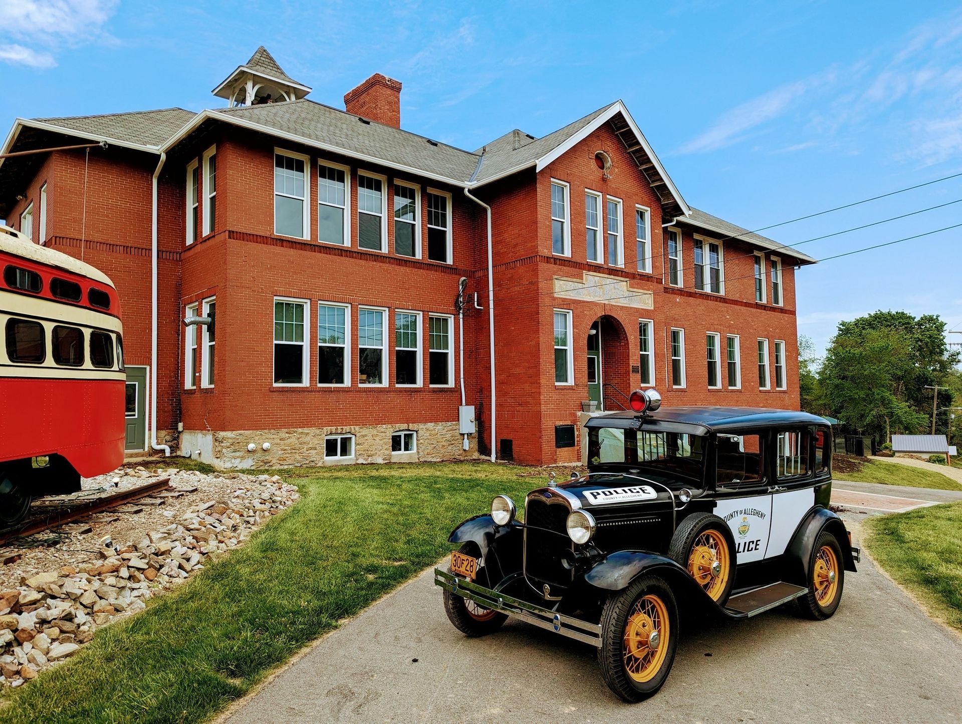 Vintage black car parked outside a large red brick building with a red trolley on the left.