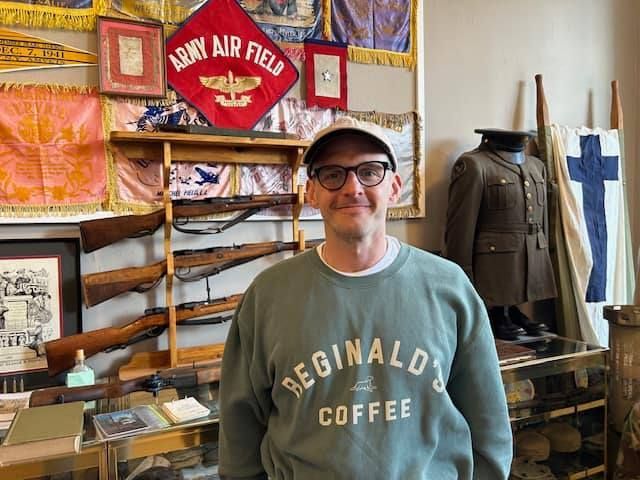 Man standing in a museum exhibit with military artifacts and a red “Army Air Field” sign behind him