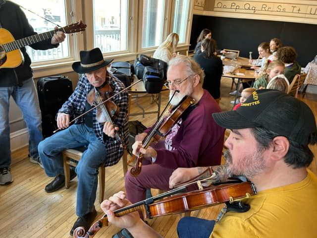 Musicians playing fiddles and guitar in a cozy wooden room with seated listeners