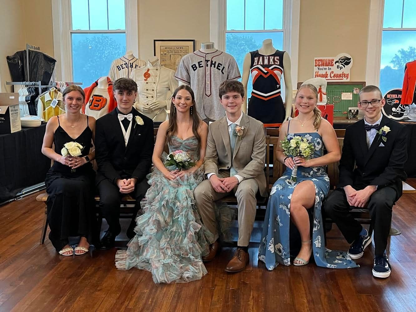 Six teens seated in formal wear at a homecoming dance, posing indoors with decorations behind them.