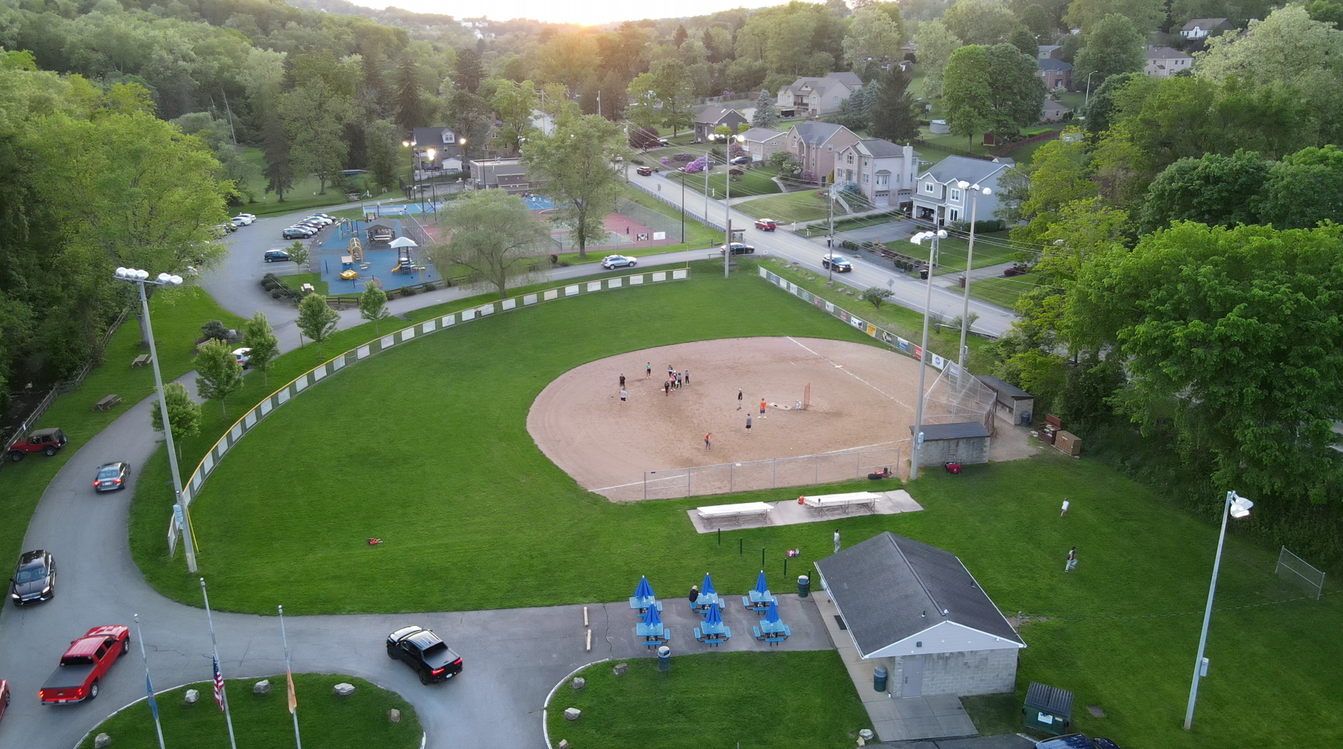 Aerial view of a park with a baseball field, green lawn, playground, and winding paths at sunset