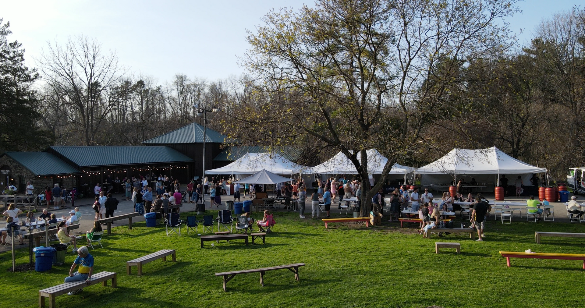 Outdoor picnic area with people gathered under white tents on a sunny day, surrounded by trees and green grass