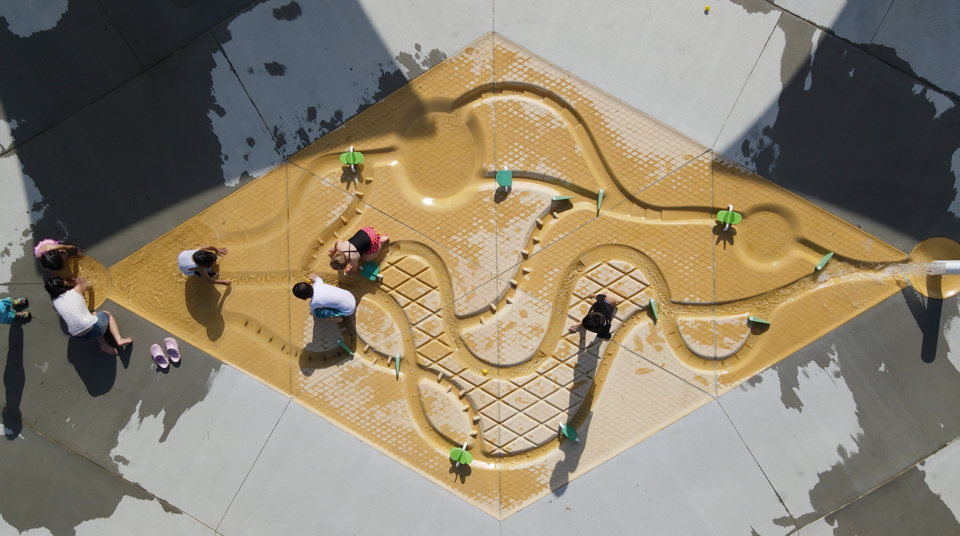 People gathered around a tan tabletop maze game outdoors, moving pieces along winding tracks.