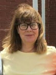 Portrait of a person with glasses and shoulder-length hair, smiling in front of a brick wall.