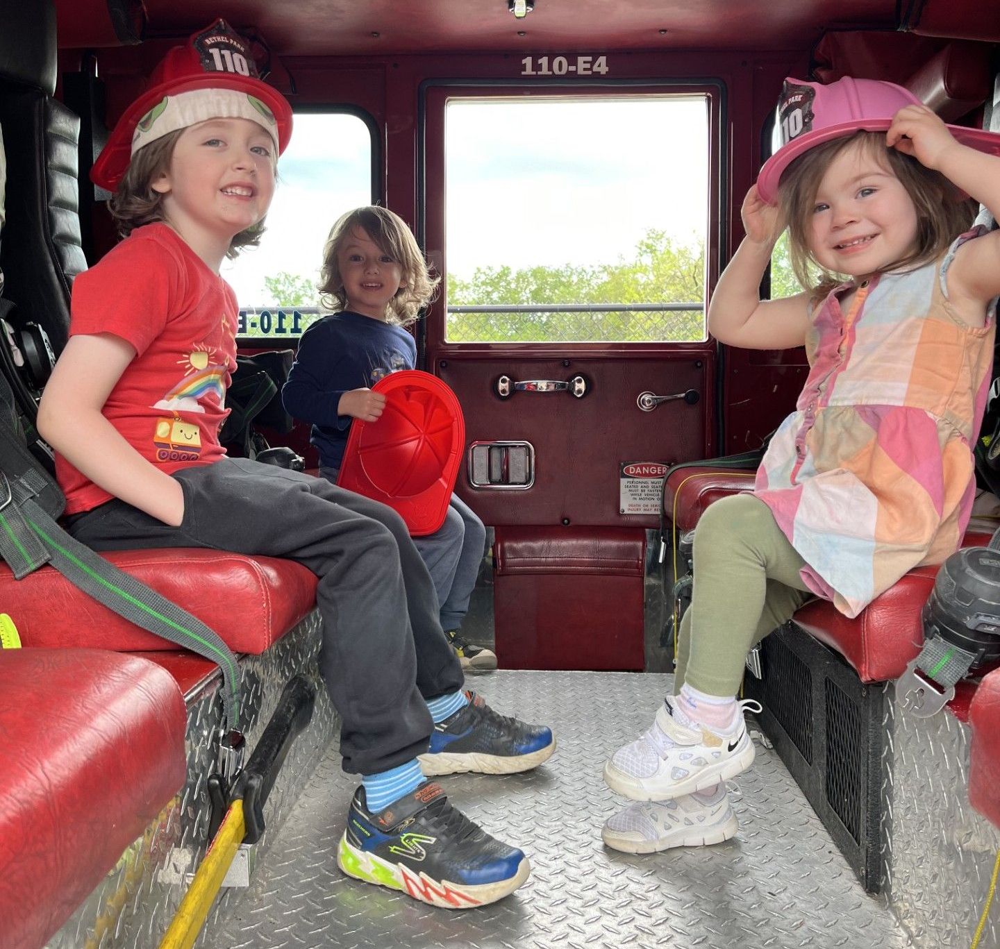 Three children sitting inside a red vehicle, smiling and wearing hats and colorful clothes.