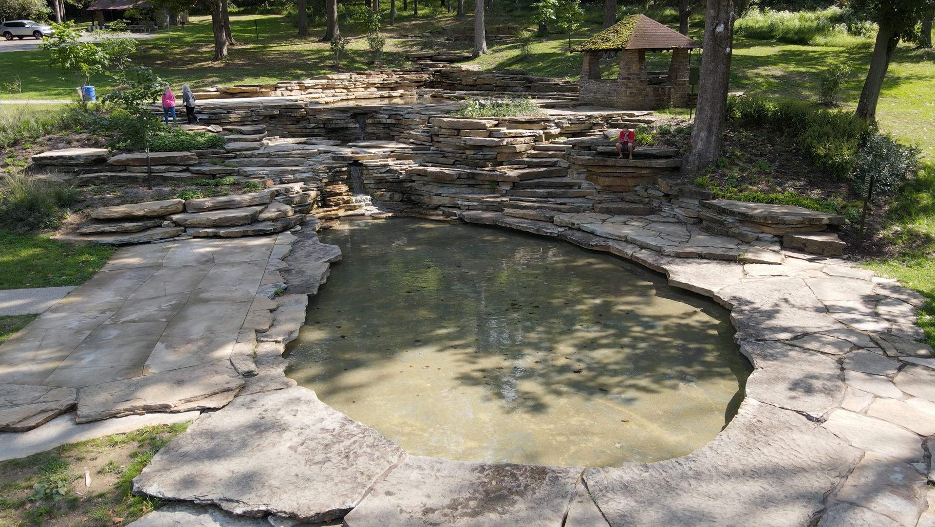 Stone-lined pond in a wooded park with steps, benches, and a small shelter nearby.