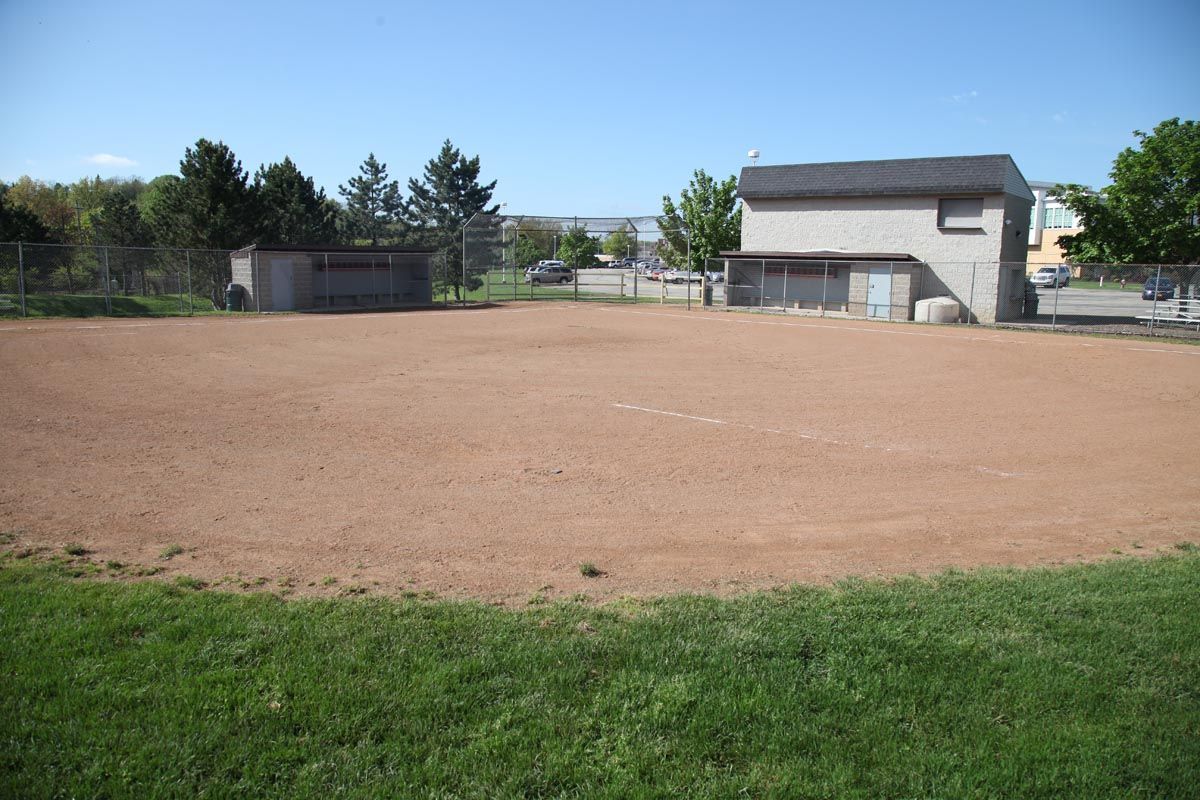 Empty dirt lot with a small building, trees, and green grass under a clear blue sky