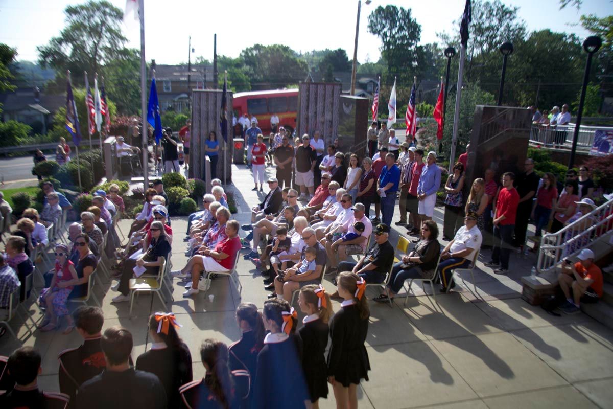Outdoor gathering of people sitting and standing in a sunlit courtyard near a stage with flags and banners