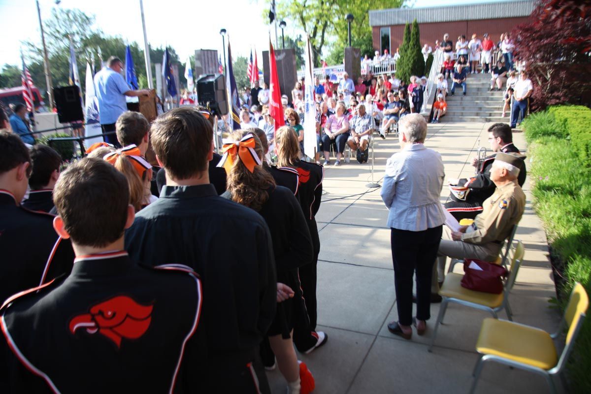 Crowd gathered outdoors on steps and sidewalk, watching a speaker near flags and a red building.