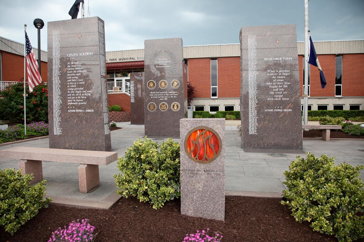 Stone memorial with engraved panels and benches in front of a brick building, surrounded by flowers and shrubs