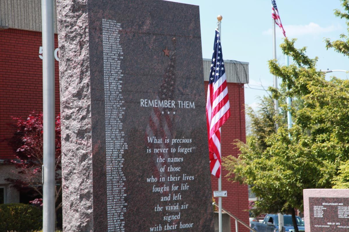 Memorial wall with engraved names and poem, beside an American flag outside a brick building.