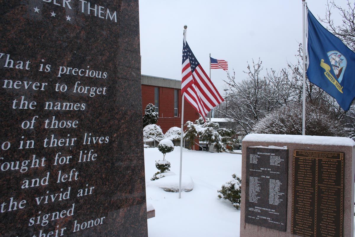 Snowy memorial with engraved stone plaques and U.S. and military flags flying beside it