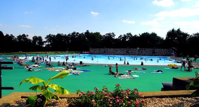 Crowded outdoor water park pool with swimmers, bright blue water, green deck, and trees in the background