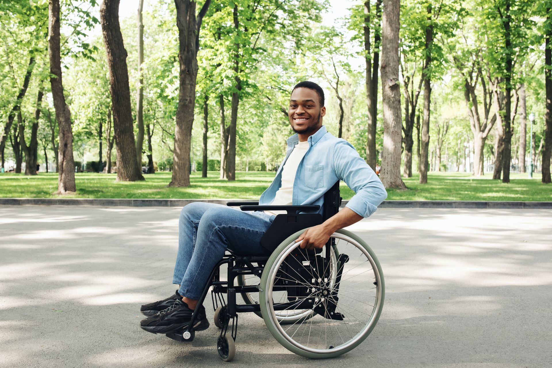 Man smiling in a wheelchair on a park path, surrounded by trees and greenery.