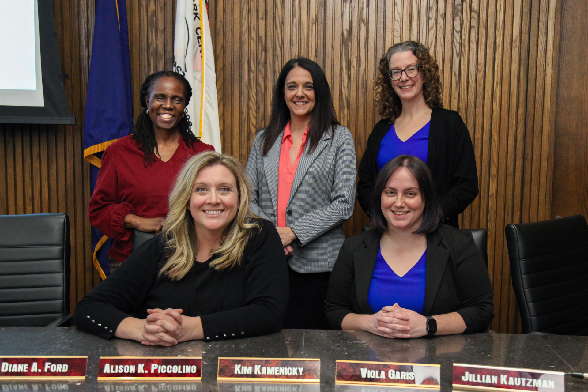 Five women pose behind and in front of a conference table in a wood-paneled meeting room.