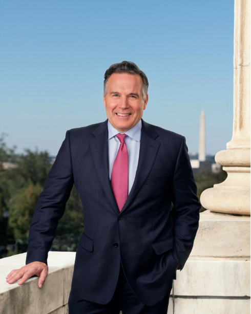 Smiling man in navy suit with pink tie standing by a stone column overlooking the Washington Monument.