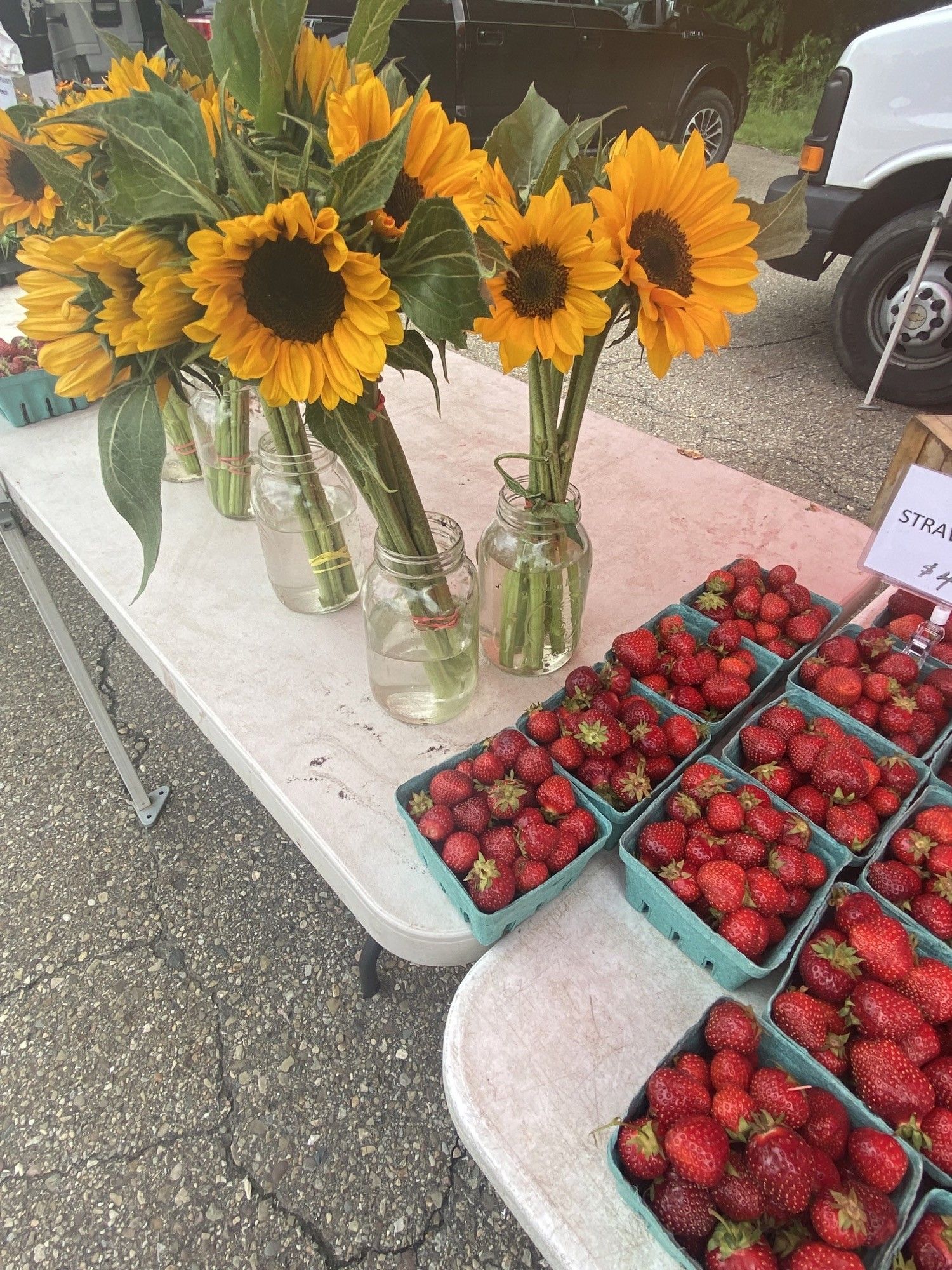 Sunflowers in glass jars beside trays of fresh strawberries at an outdoor market table