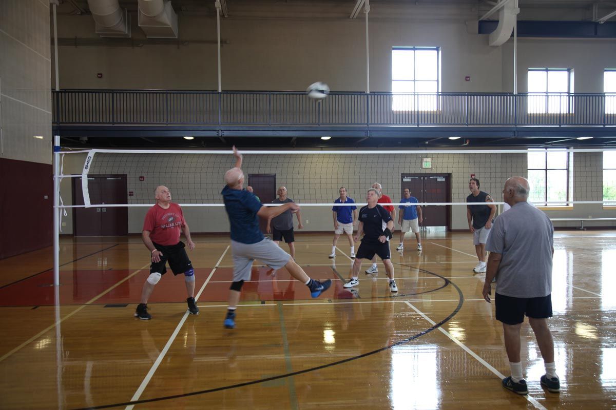 Volleyball game in a gym, with players jumping and reaching near the net on a polished court.