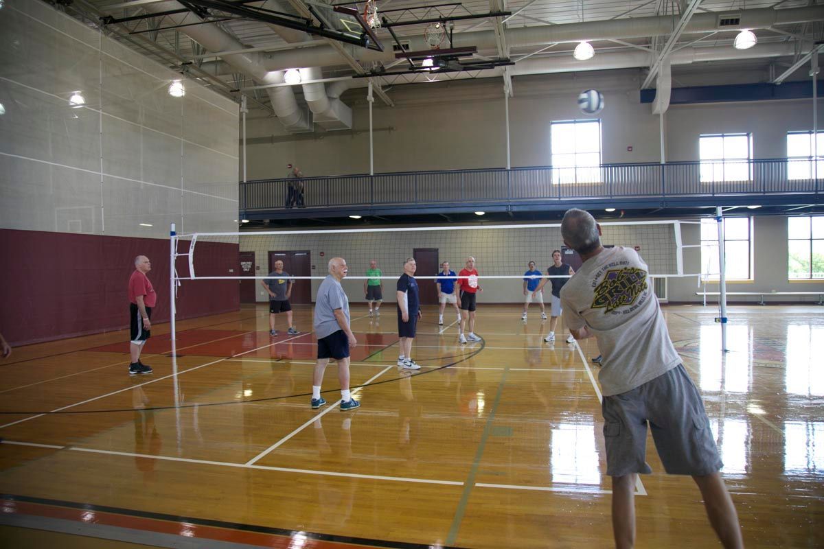 People playing volleyball in an indoor gym, with one player serving in the foreground