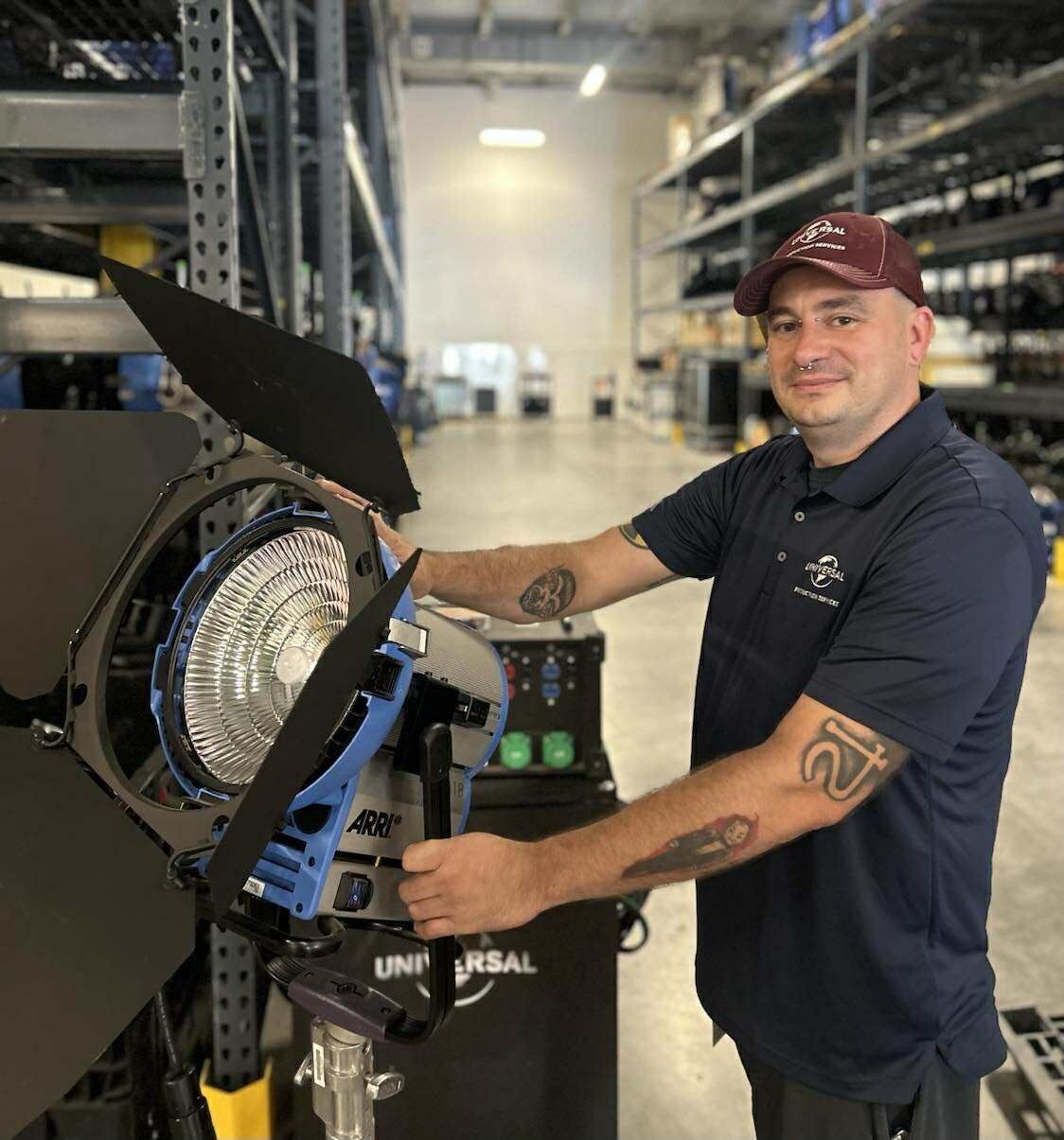 Man in a warehouse holds a studio light on a stand, smiling in a warehouse aisle.