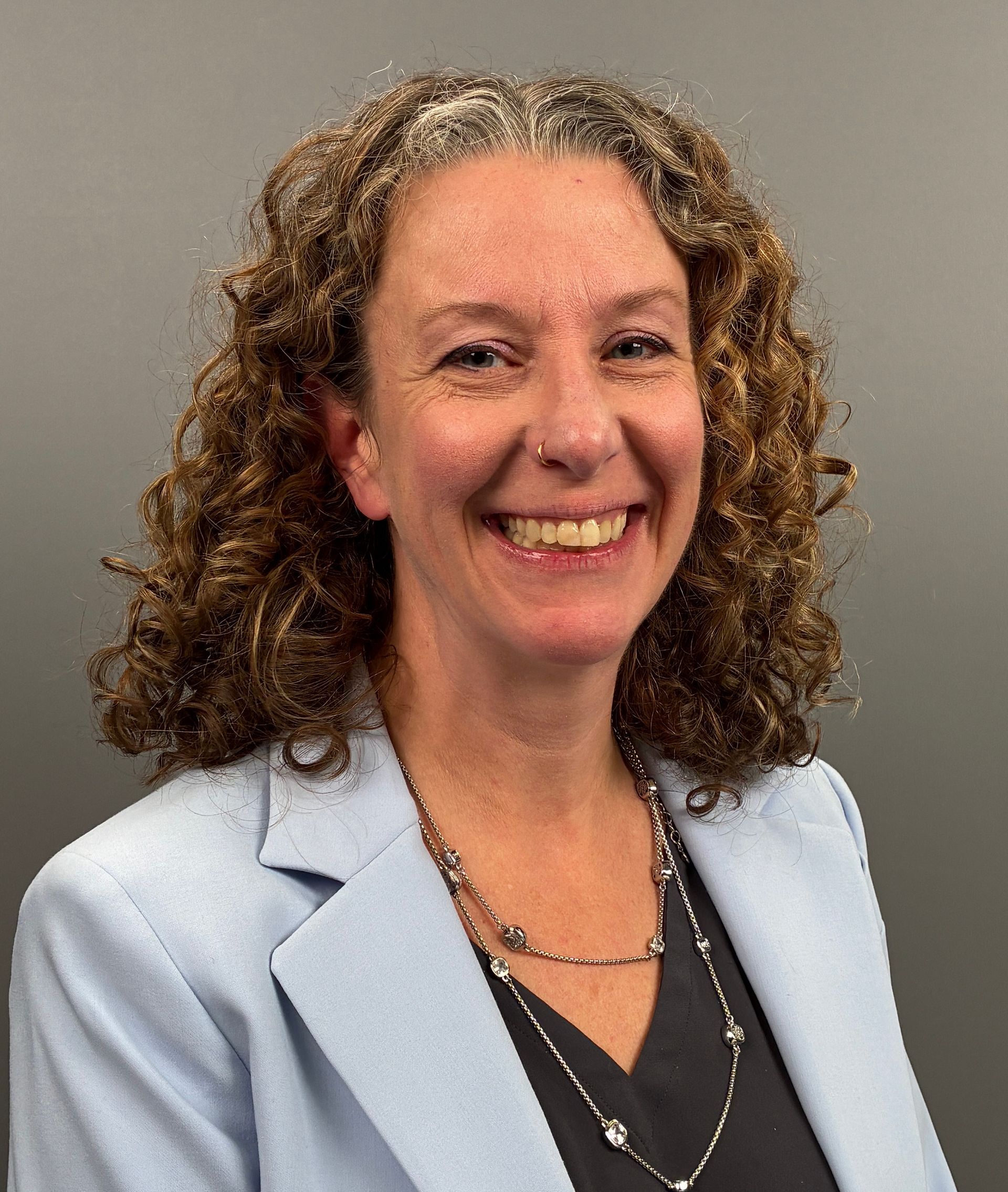 Smiling person with curly hair in a light blue blazer against a gray background