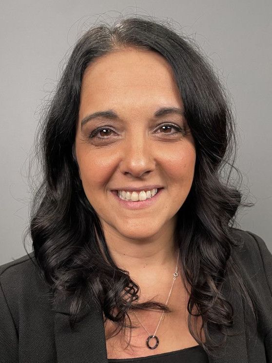 Portrait of a smiling person with dark wavy hair against a plain gray background