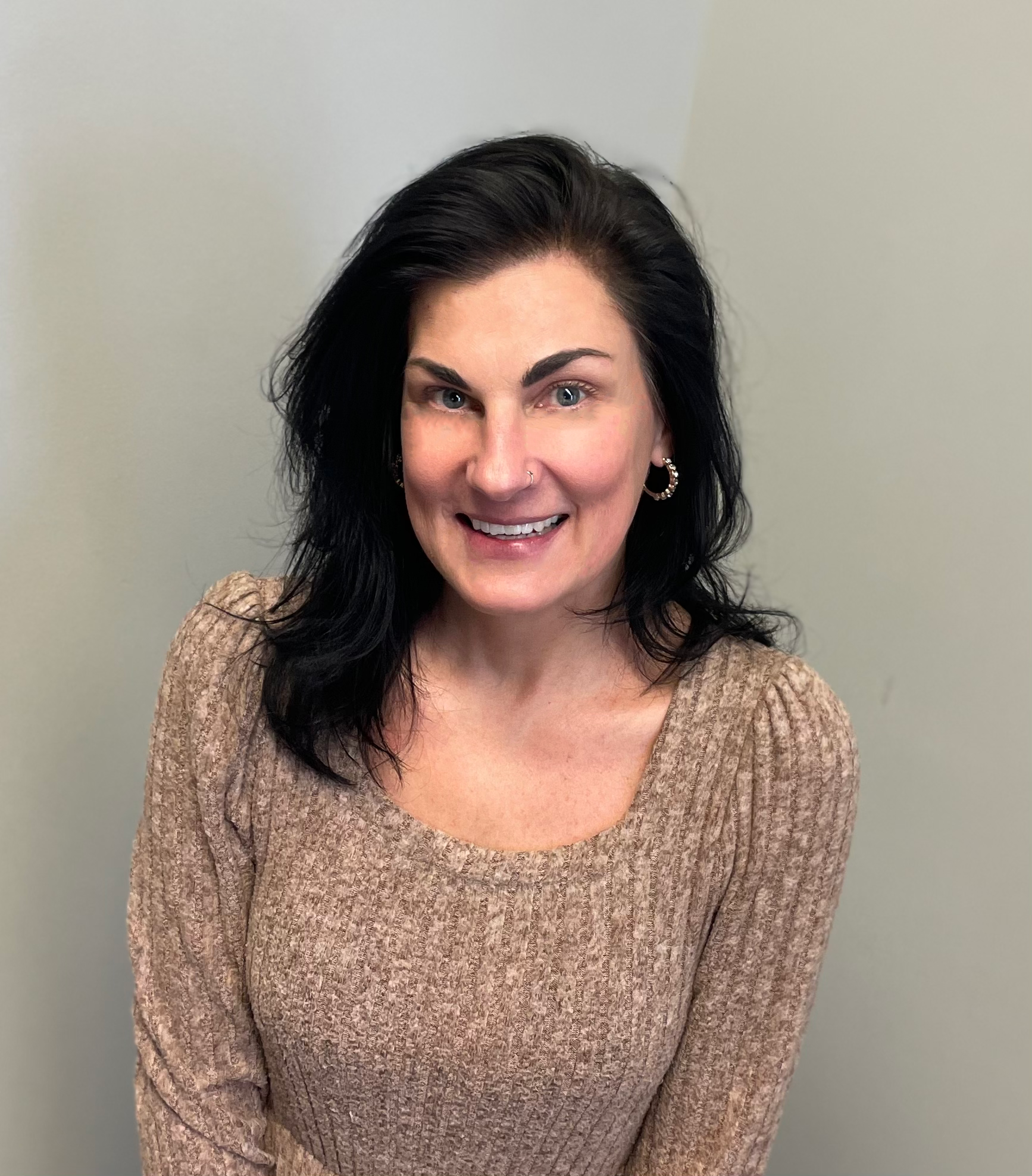 Smiling woman with dark hair wearing a beige sweater against a light gray background