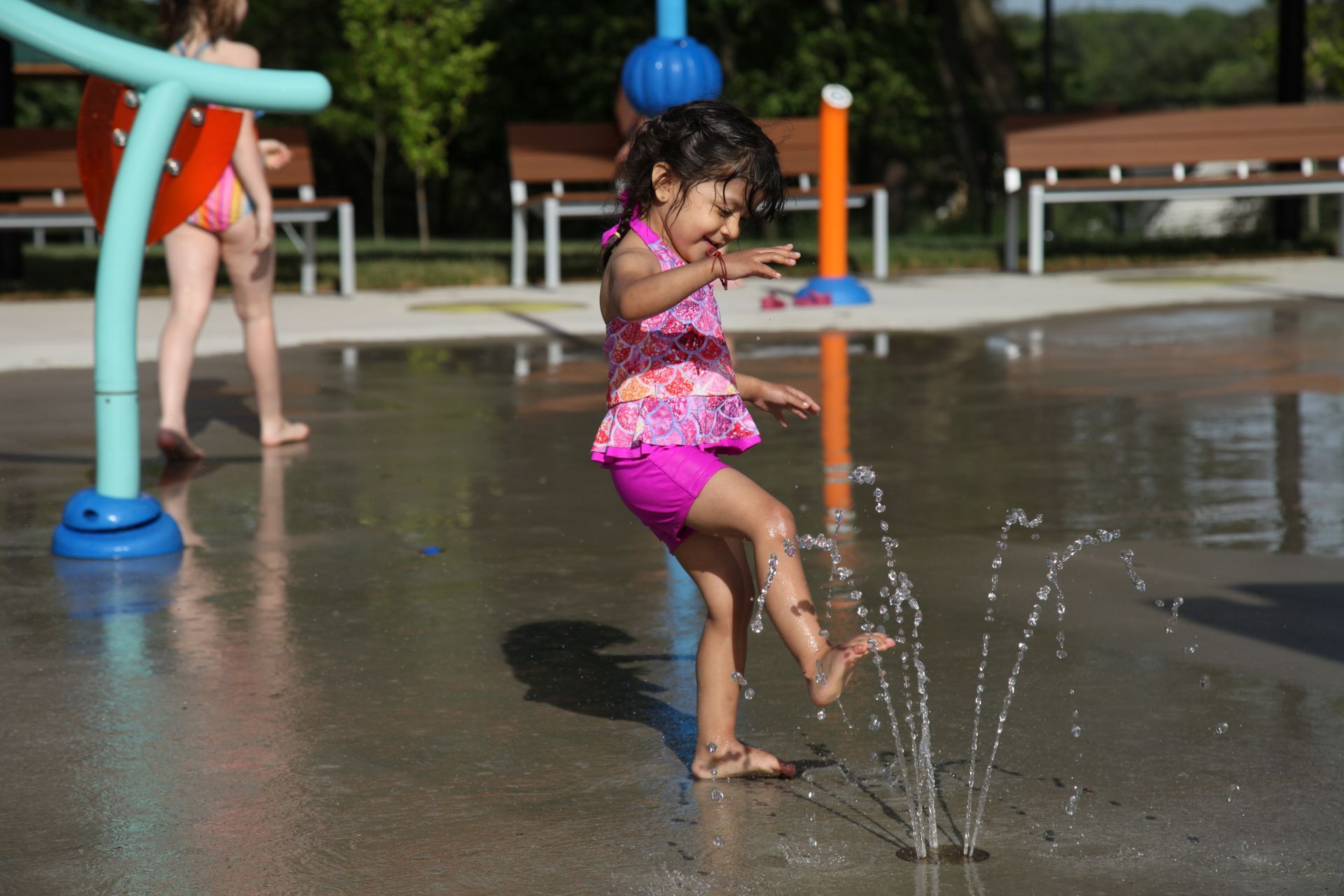 Child in pink swimwear splashes water at an outdoor spray park on a sunny day