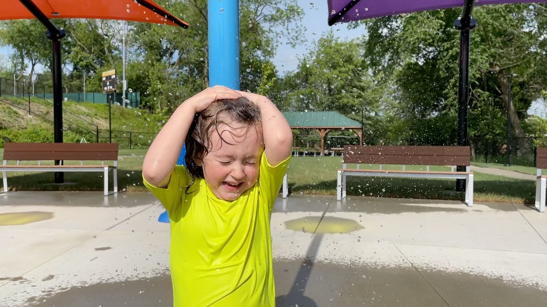Child in a bright yellow shirt under a splash pad, holding head as water sprays in a park
