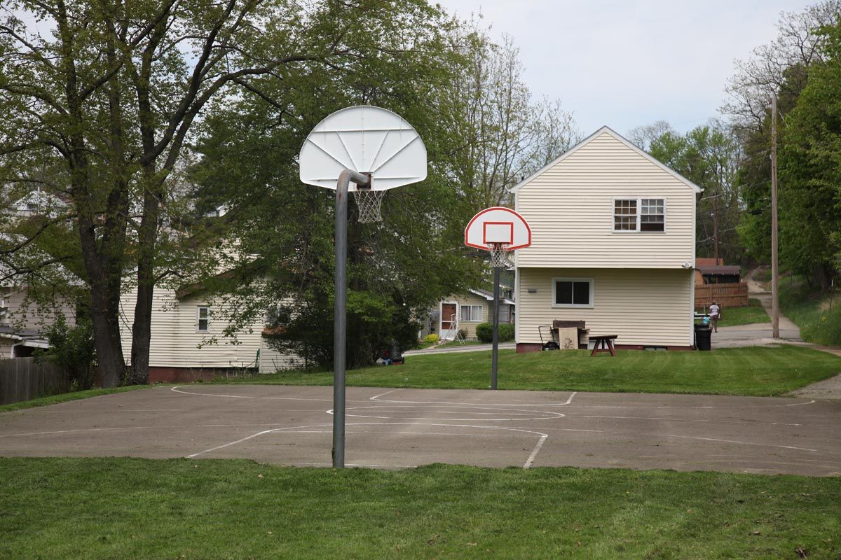 Outdoor basketball court with two hoops beside a white house and trees.