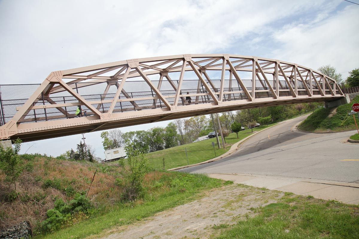 Rusty metal truss bridge spanning over a road beside a grassy embankment under a cloudy sky