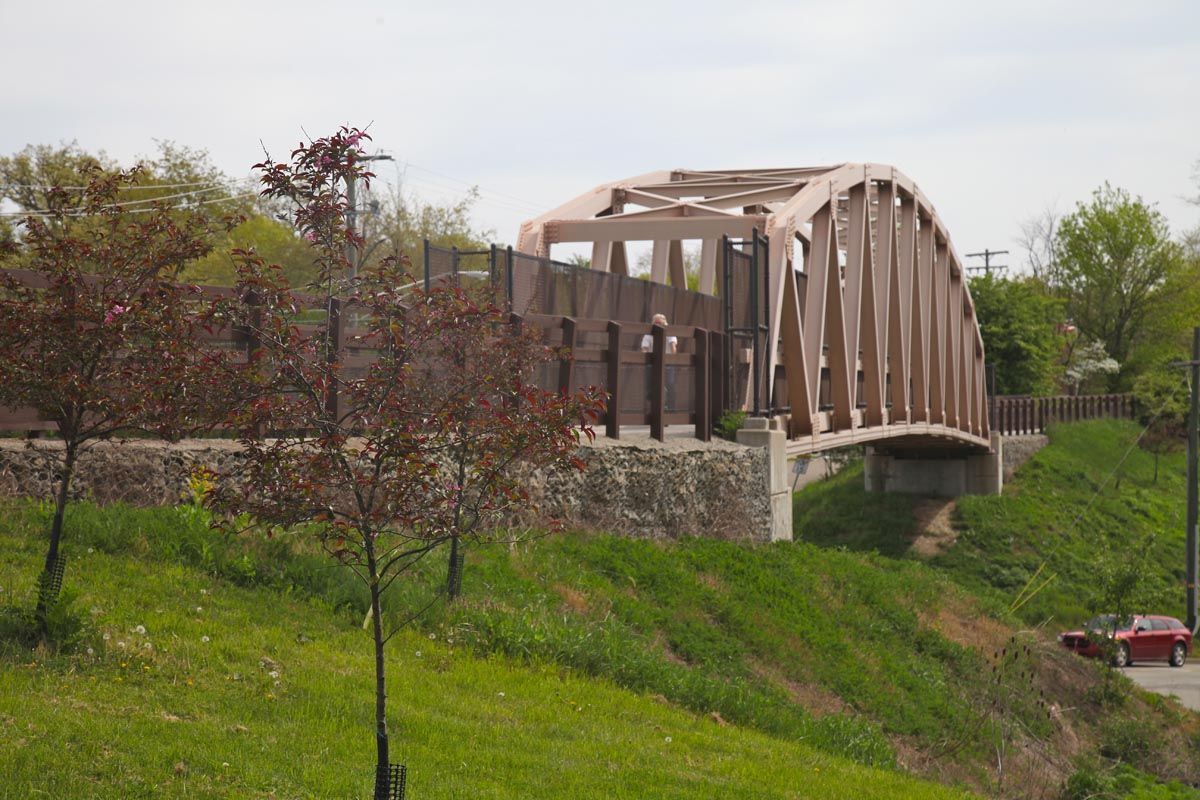 Arched steel bridge over a grassy slope, with trees and a small red car below.