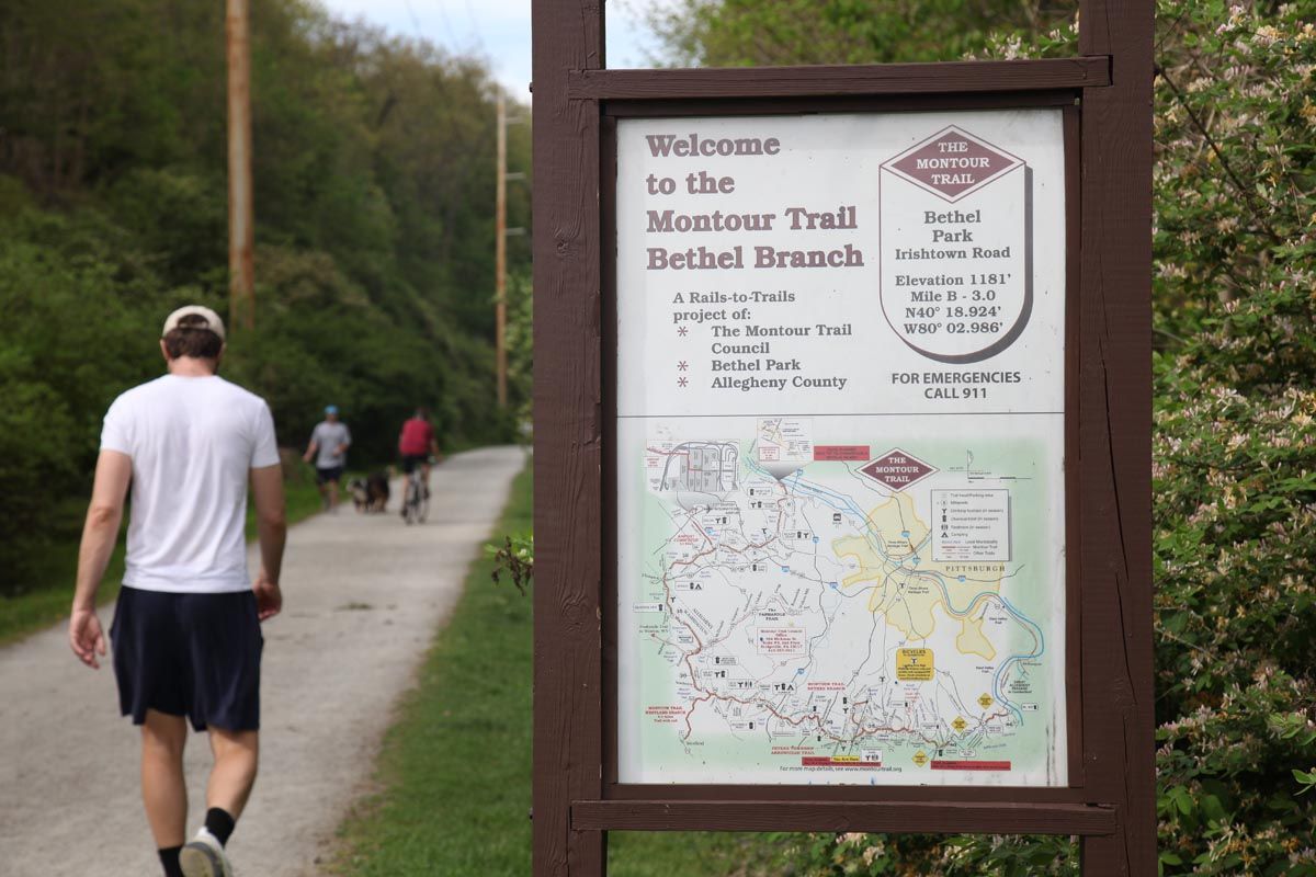 Trail entrance sign with map beside a wooded path, with two people walking away