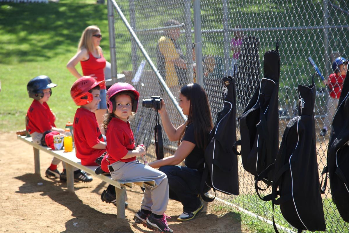 Children in red uniforms sitting on a bench by a chain-link fence at a sports field