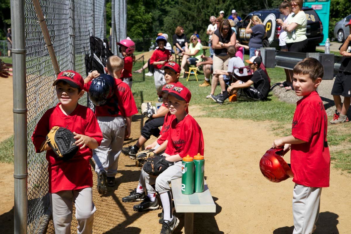 Children in red shirts and baseball uniforms standing near a dugout at a sunny ball field.