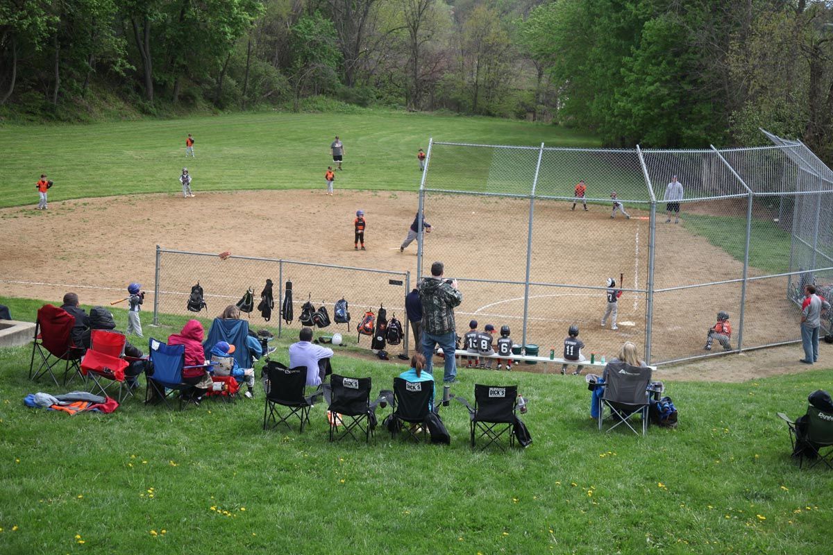 People seated near a softball field behind a chain-link backstop, watching a game on a grassy hillside.