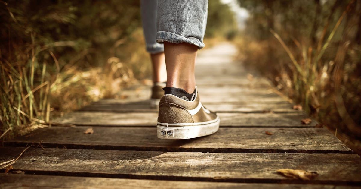 Person walking on a wooden path in sneakers and rolled-up jeans, with sunlight and dry grass nearby