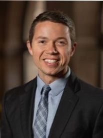 Smiling man in a dark suit and blue tie against a blurred indoor background
