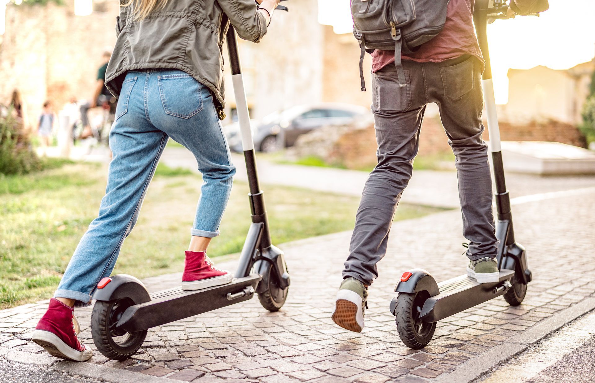 Two people riding electric scooters on a sunlit cobblestone street