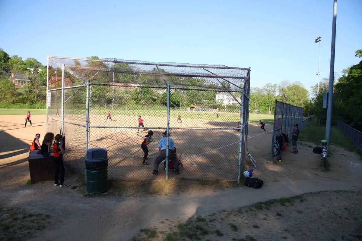 Baseball field with players near a batting cage, seen through chain-link fencing on a sunny day