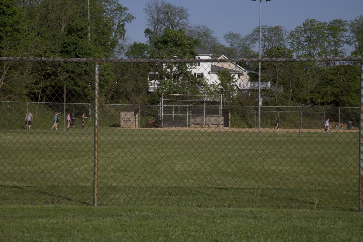 A chain-link fence in front of a grassy field with a few people and a house in the background.