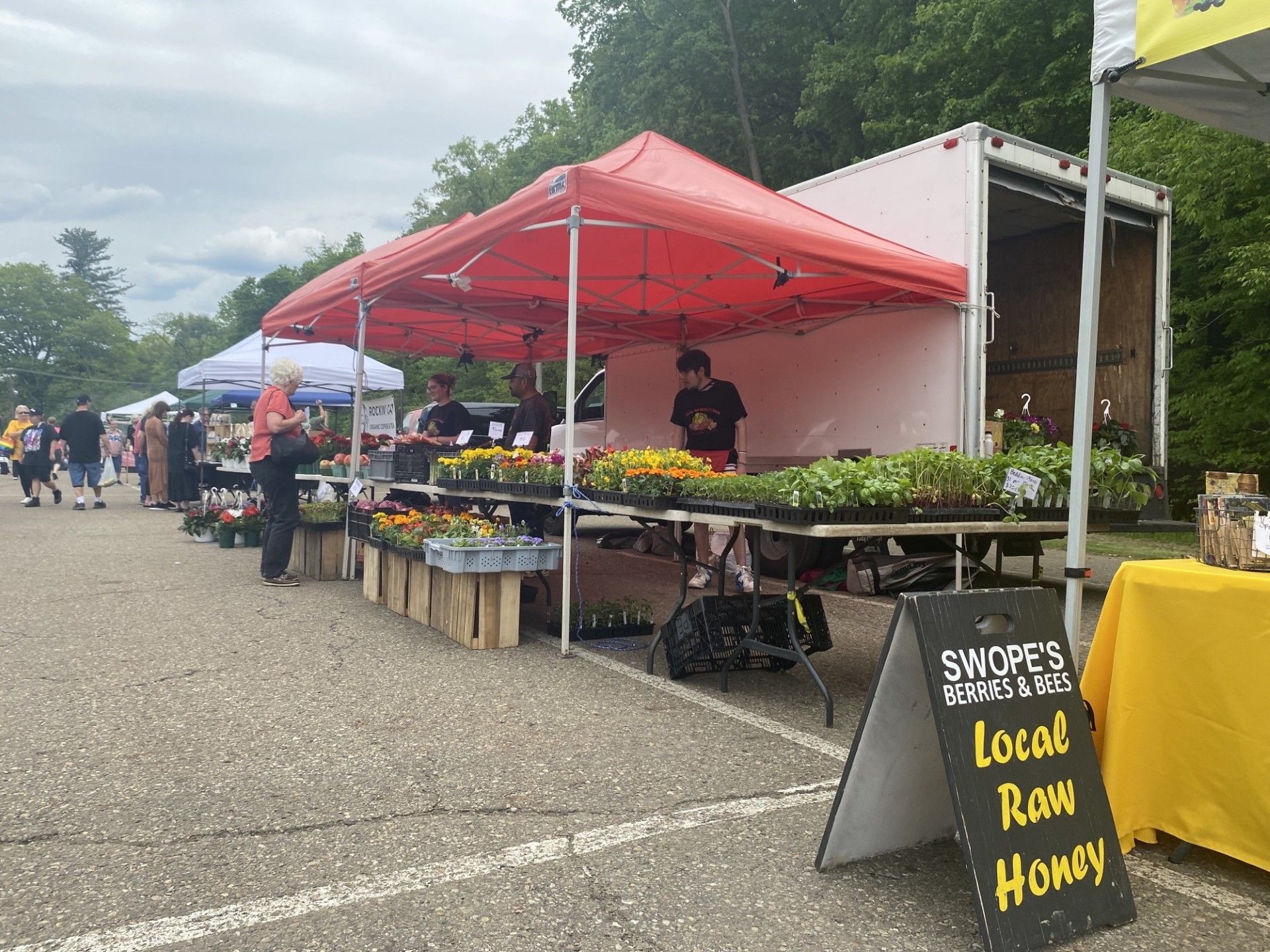 Outdoor local farm market stalls under red tents with produce and a sign reading “Local Fesh Honey.”