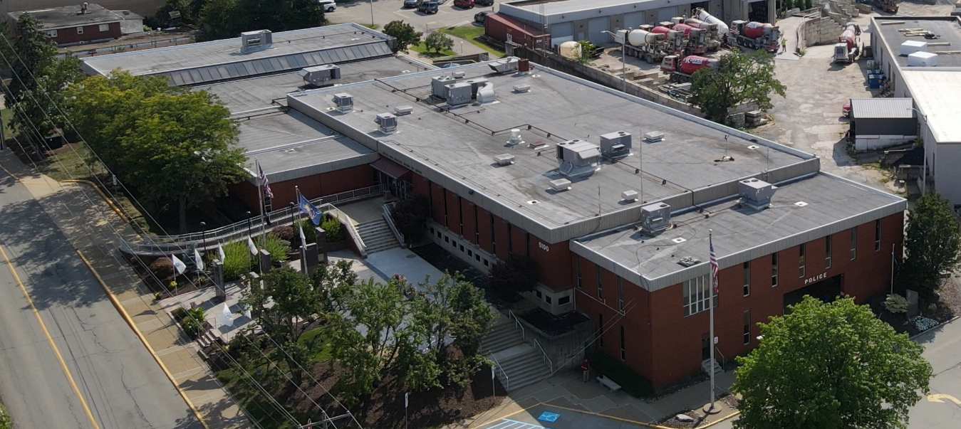 Aerial view of a brick school building with rooftop HVAC units and parking lots surrounded by trees