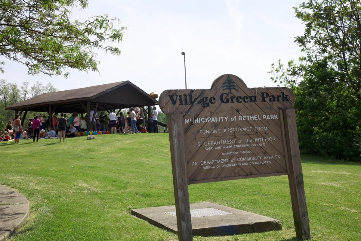 Village Green Park sign in front of a pavilion with people gathered on a grassy lawn.